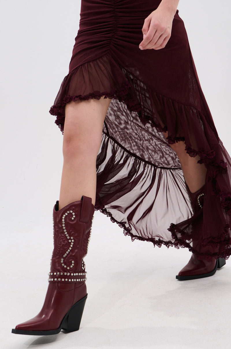A person walking forward on a white background wears a burgundy high-low ruffled dress with baroque embroidery and the AZALEA WANG UMBRA BURGUNDY WESTERN BOOTIE adorned with silver studs.