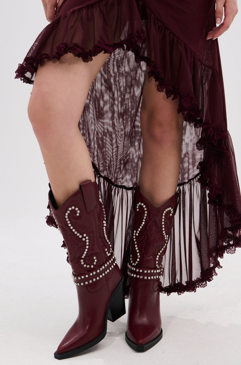 A person wearing a burgundy high-low ruffled dress and AZALEA WANG UMBRA BURGUNDY WESTERN BOOTIE with decorative stitching, Baroque embroidery, and silver studs, standing on a white background.