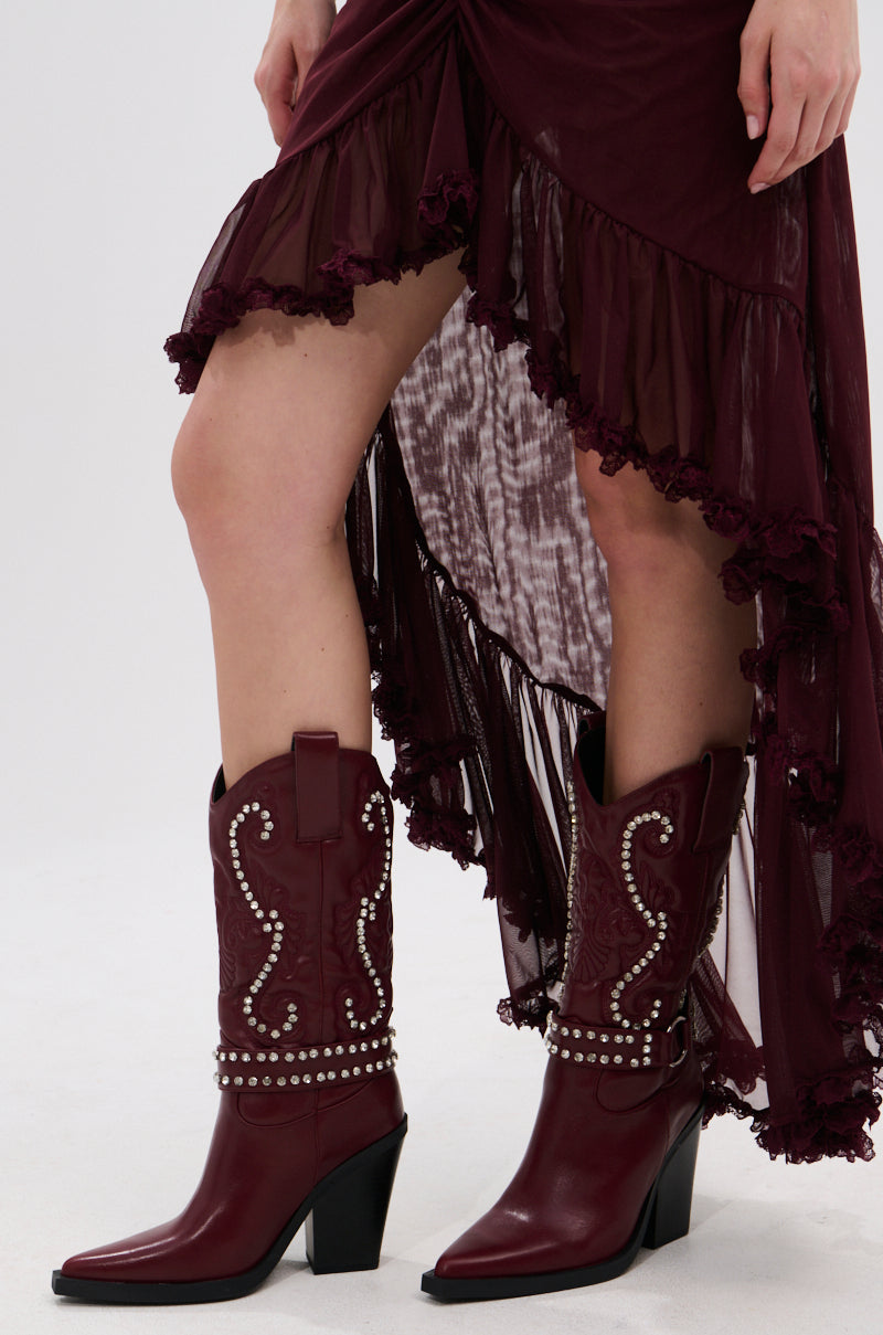 A person wears a burgundy high-low ruffled skirt and AZALEA WANG UMBRA BURGUNDY WESTERN BOOTIE with silver studs and baroque embroidery, standing on a white background.