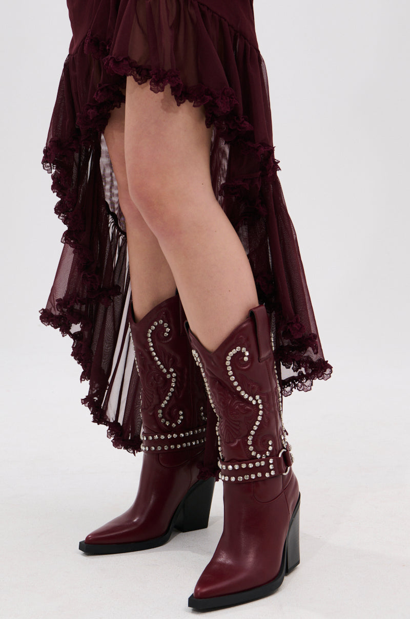 A person wearing a burgundy sheer ruffled high-low skirt and AZALEA WANG UMBRA BURGUNDY WESTERN BOOTIE with Baroque embroidery and decorative studs, standing on a light background.