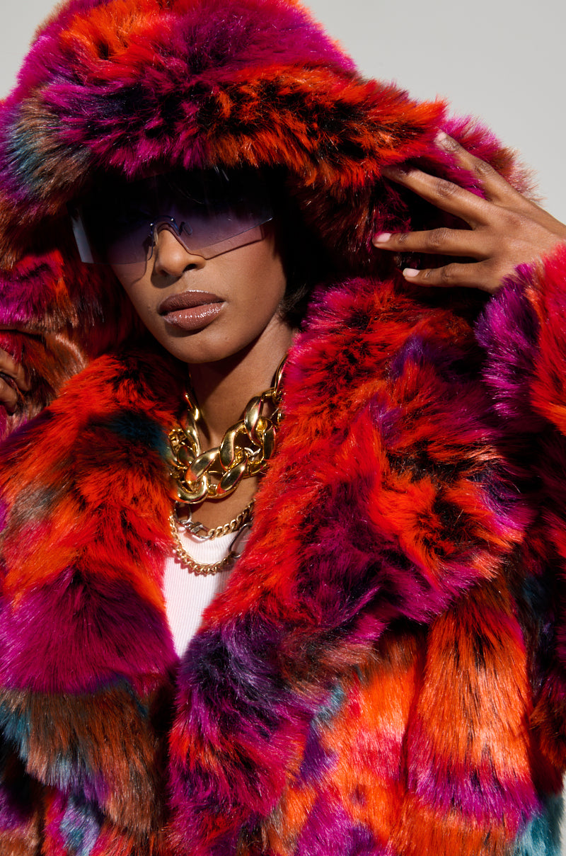 A person dons the UPSETTER MAXI FAUX FUR COAT IN PINK MULTI featuring an oversized design with a large hood, paired with sleek sunglasses and a chunky gold necklace. With their hands adjusting the hood, they gaze ahead confidently, exuding an air of bold sophistication.