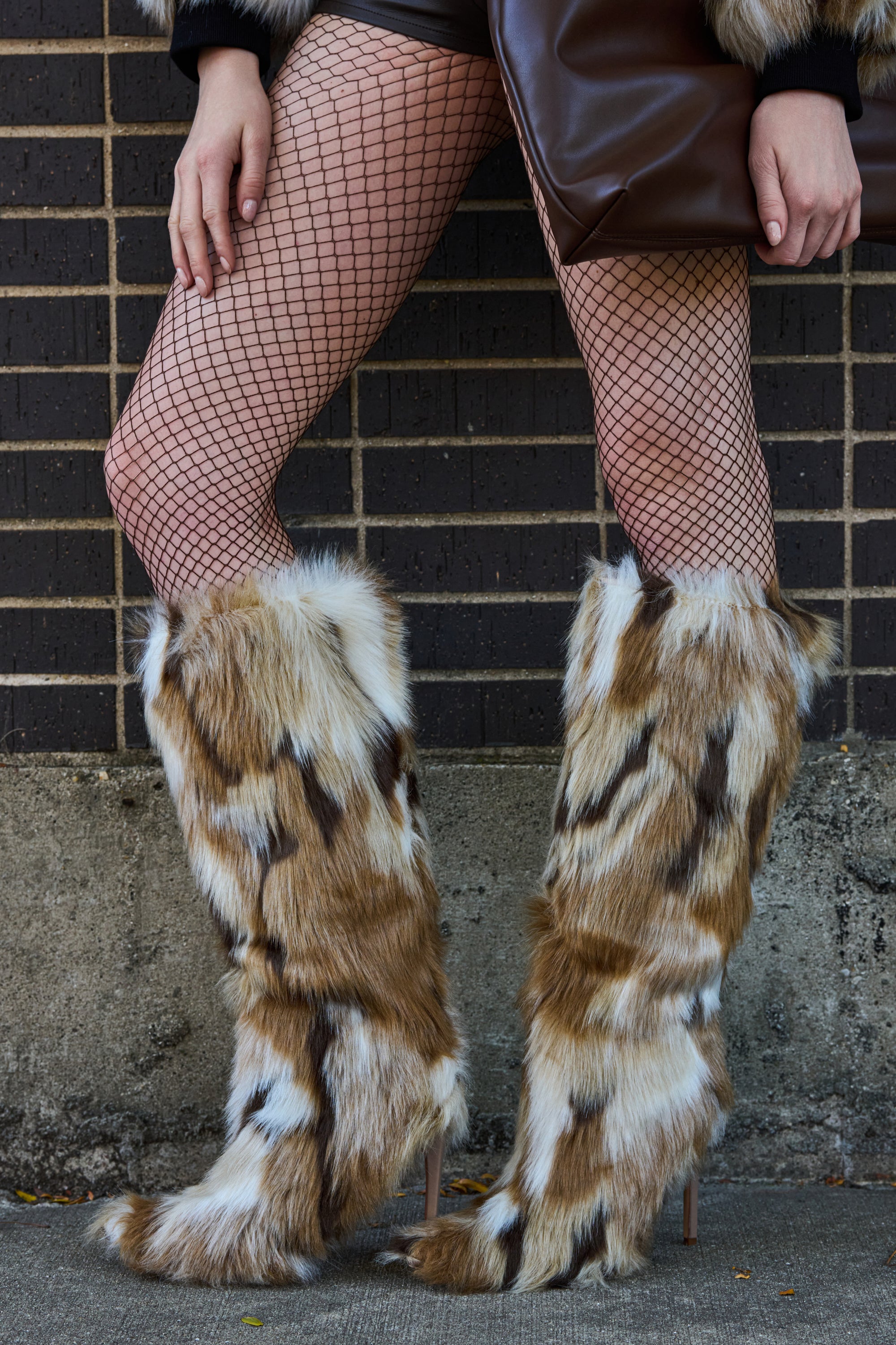 A person stands against a brick wall, wearing fishnet stockings and the AZALEA WANG UPSETTER MULTI FURRY BOOT with pointed toe and stiletto heel. They hold a large brown clutch bag, with only their legs and hands visible.