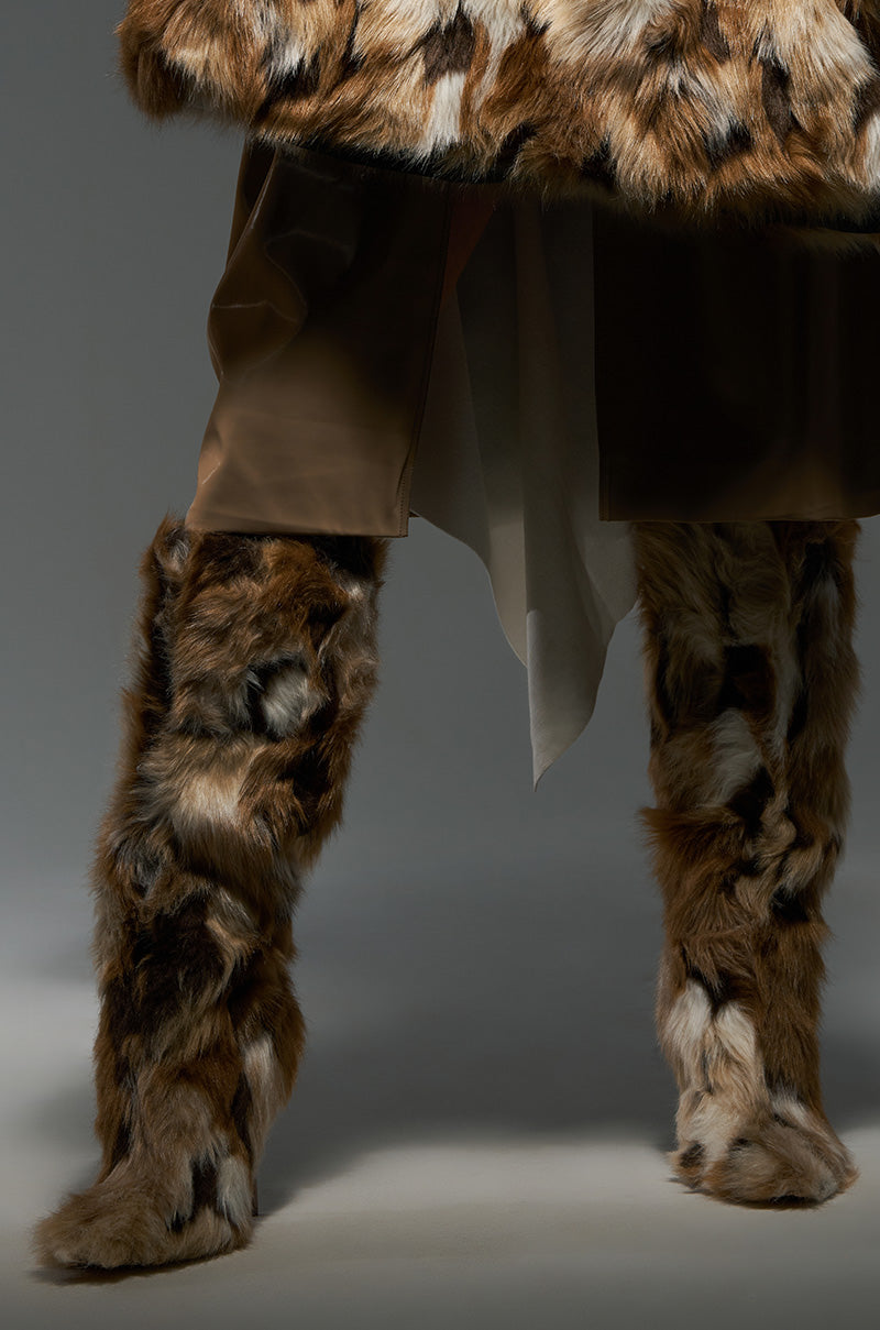 A person is wearing a fur coat with matching AZALEA WANG UPSETTER MULTI FURRY BOOTS in a brown, black, and white pattern. Standing against a neutral backdrop, they sport dark pants or a skirt visible between the coat and their pointed toe boots.