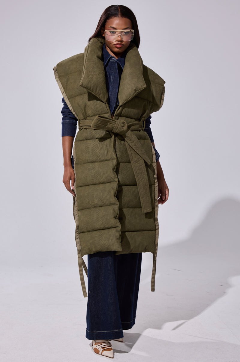 A woman styles the PILOT JONES LONGLINE PUFFER VEST IN CROC SUEDE over a dark denim shirt and flared jeans, accessorizing with clear-framed glasses and white pointed-toe shoes.