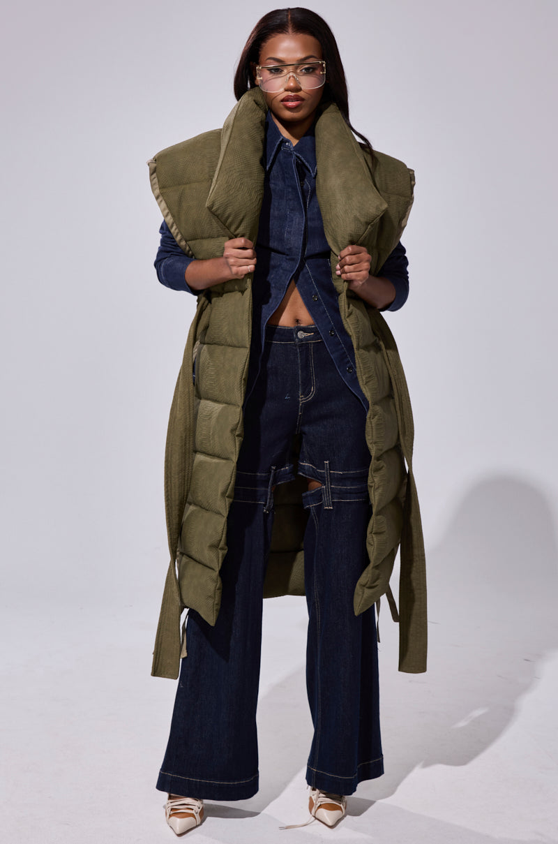 A woman wears the PILOT JONES LONGLINE PUFFER VEST IN CROC SUEDE over a dark denim button-up shirt and wide-leg jeans, styled with clear glasses and pointed cream heels against a plain, light background.
