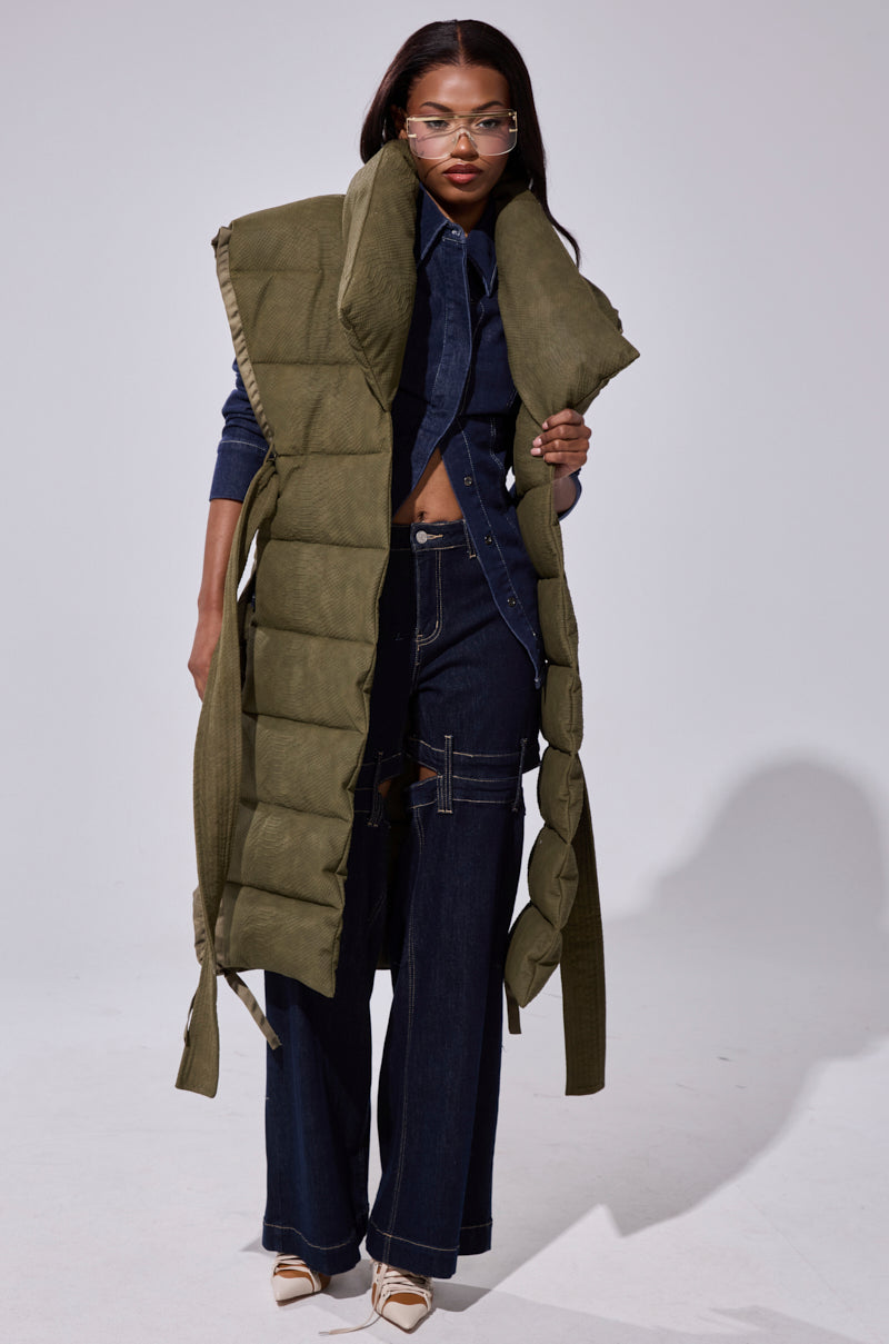 A woman wearing the PILOT JONES LONGLINE PUFFER VEST IN CROC SUEDE over a dark denim jumpsuit with white stitching and wide legs, paired with clear glasses and cream heels, stands against a plain light background.
