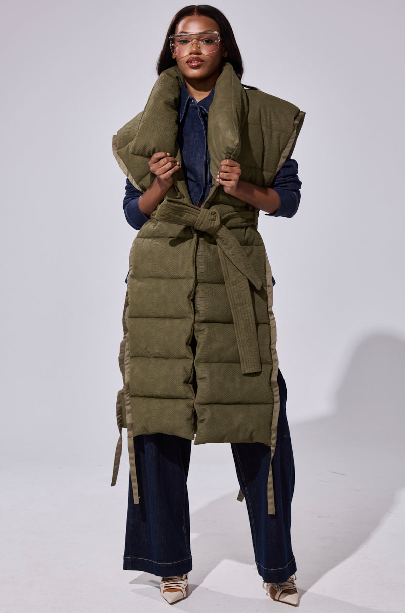 A woman models the PILOT JONES LONGLINE PUFFER VEST IN CROC SUEDE, featuring an oversized collar and belted waist, layered over dark denim and pointed-toe shoes. She finishes her look with clear protective glasses.
