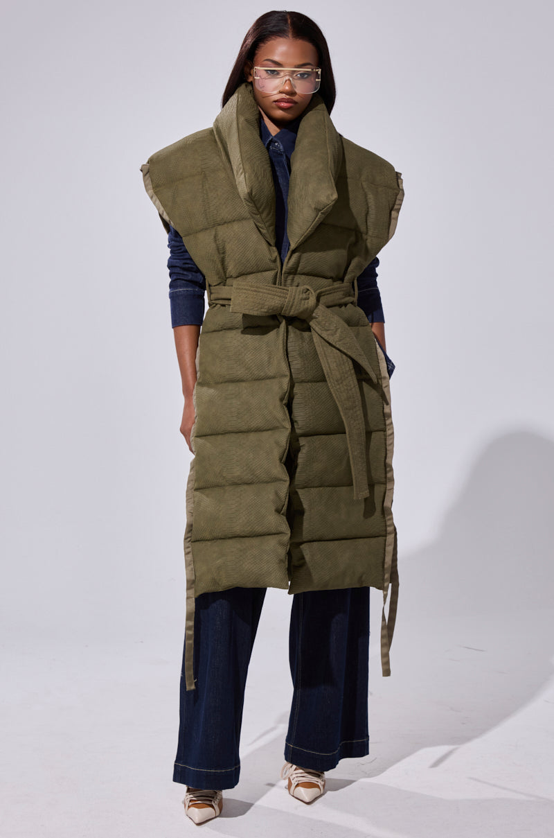 A woman wearing glasses models the PILOT JONES LONGLINE PUFFER VEST IN CROC SUEDE with an oversized collar in olive green, belted at the waist over dark denim and cream pointed shoes, standing against a plain light background.