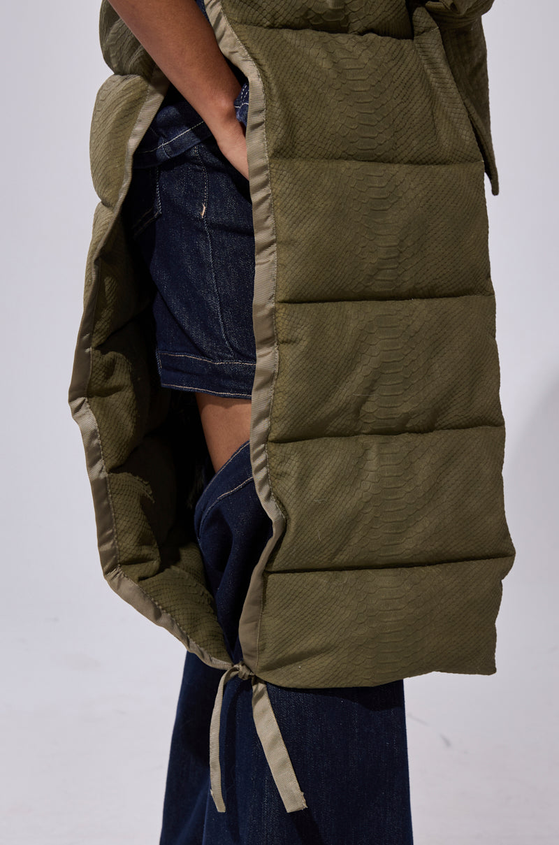 A close-up of a person wearing the PILOT JONES LONGLINE PUFFER VEST IN CROC SUEDE with side tie detail, styled open over dark denim shorts and wide-leg jeans, showing layered clothing underneath.