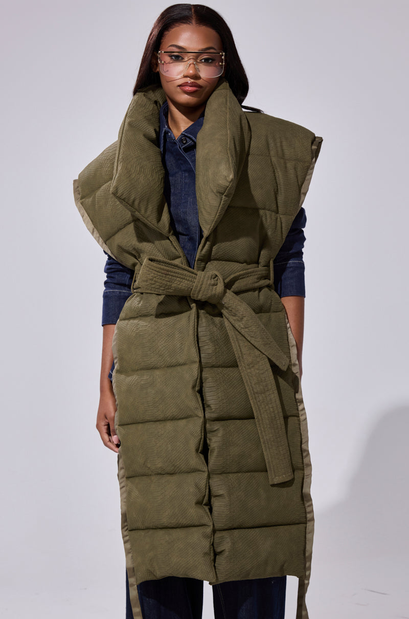 A woman in glasses wears the PILOT JONES LONGLINE PUFFER VEST IN CROC SUEDE with an oversized collar over dark denim, standing against a light background.