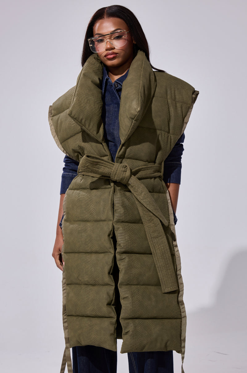 A woman models the PILOT JONES LONGLINE PUFFER VEST IN CROC SUEDE—an olive green, sleeveless vest with an oversized collar and matching belt—styled with oversized clear glasses and dark denim against a plain background.