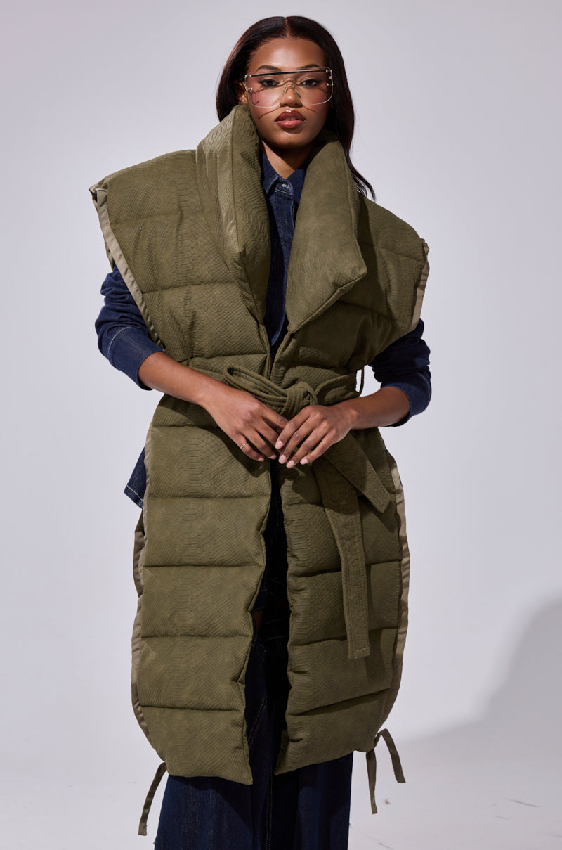 A woman with long, straight hair wears gold glasses and the PILOT JONES LONGLINE PUFFER VEST IN CROC SUEDE, belted over a dark denim outfit, standing confidently against a plain light background.