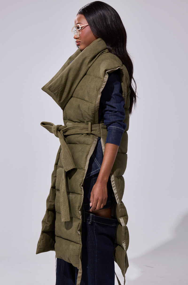 A woman in glasses and dark layered clothing models the PILOT JONES LONGLINE PUFFER VEST IN CROC SUEDE, featuring an oversized collar and belted waist, standing in profile against a plain light background.