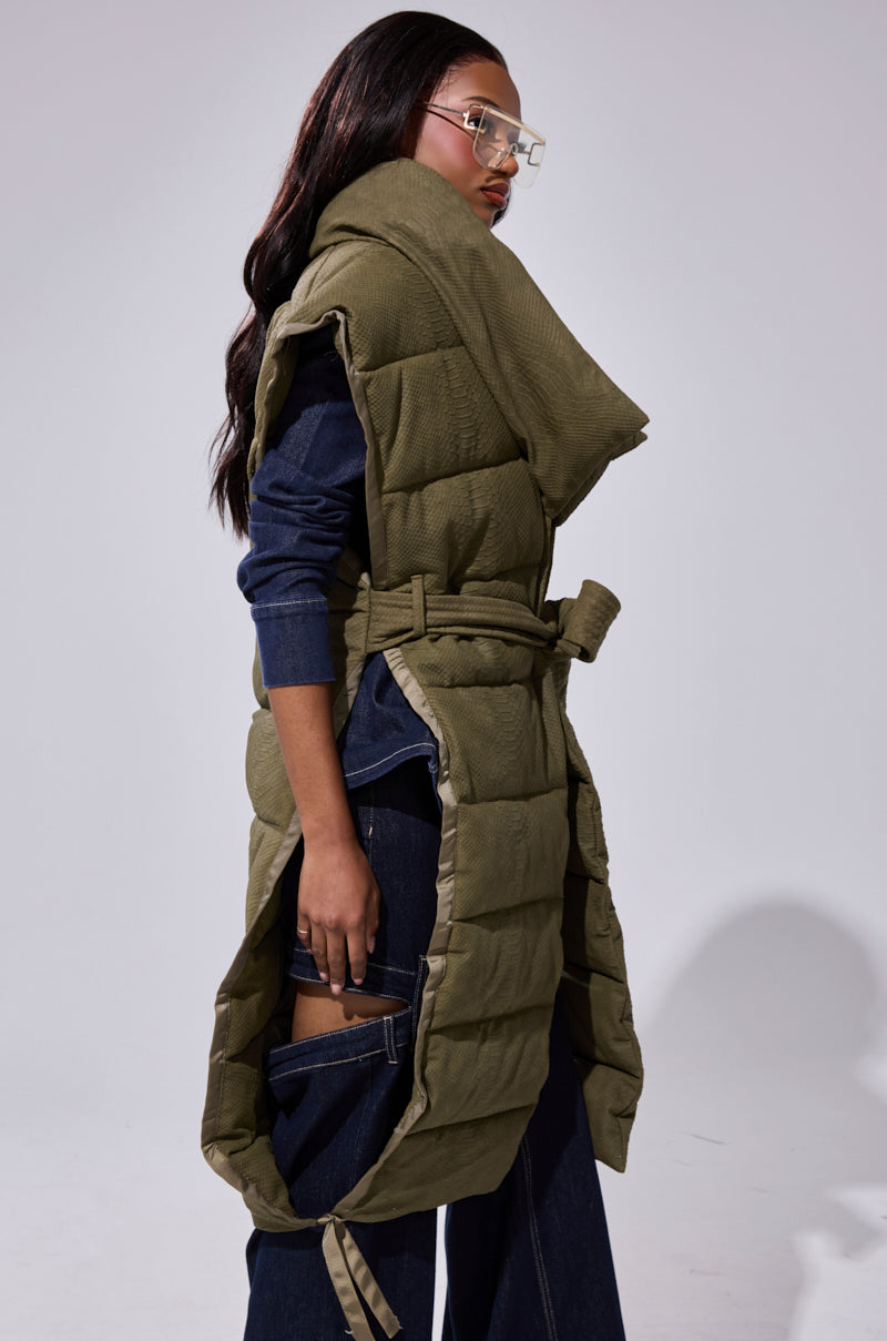 A woman in gold-rimmed glasses, the PILOT JONES LONGLINE PUFFER VEST IN CROC SUEDE with an oversized collar and belt, and dark blue wide-leg jeans with side cutouts poses against a plain light background.