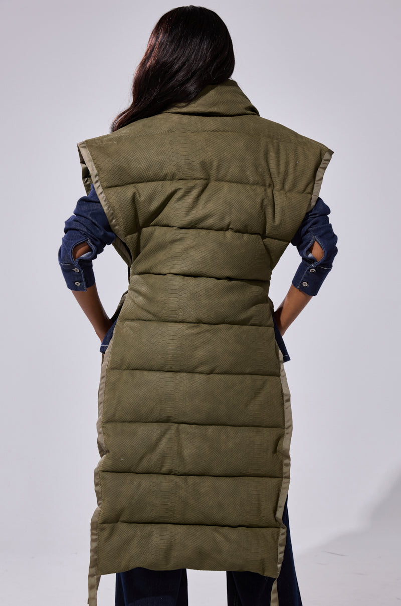 A person stands with their back to the camera, wearing the PILOT JONES Longline Puffer Vest in croc suede, an olive-green quilted design falling below the knees, layered over a dark blue long-sleeve top and dark pants.