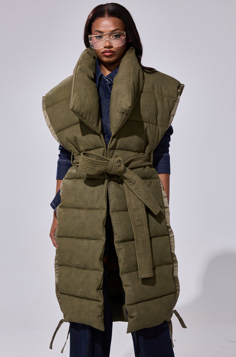 A woman wearing clear glasses and the PILOT JONES LONGLINE PUFFER VEST IN CROC SUEDE over a dark denim outfit stands against a plain light gray background.