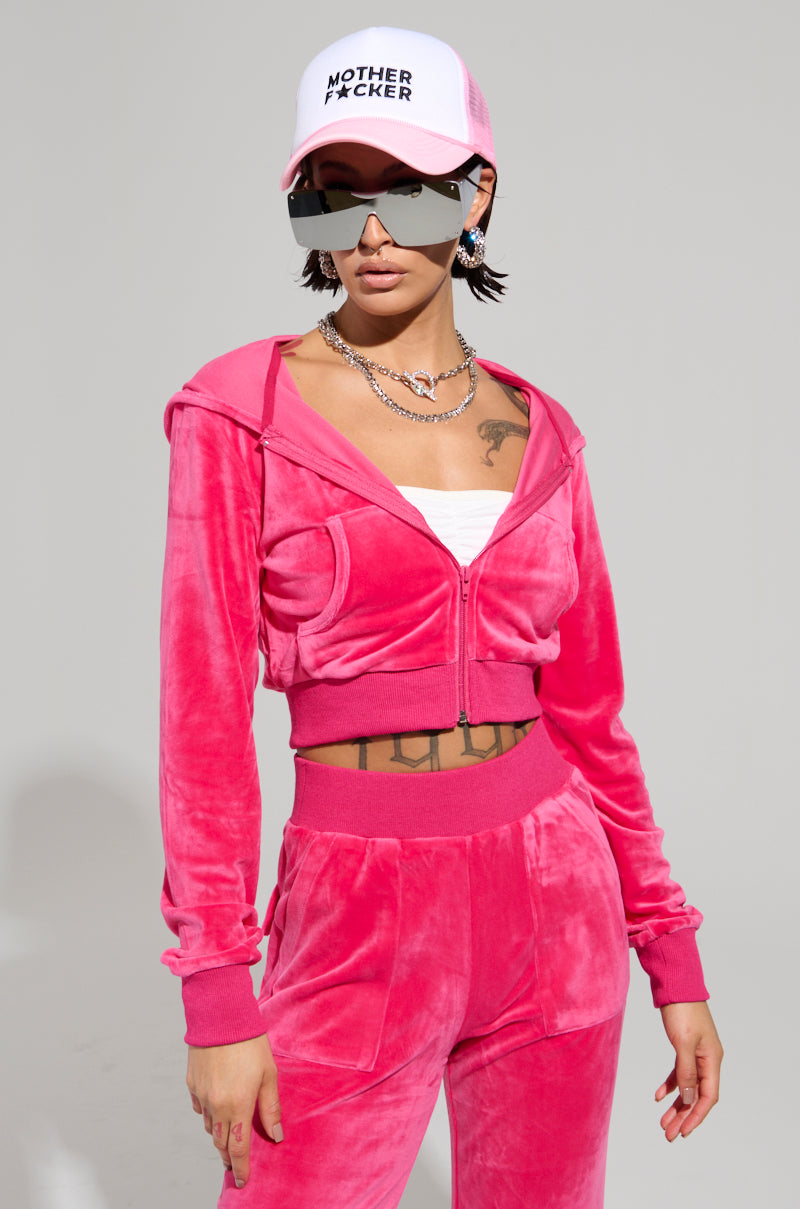 Dressed in a VELOUR GALORE FITTED ZIP UP IN PINK, a person exudes confidence with their white cap featuring black text, large sunglasses, and layered chain necklaces. Their short hair exposes Y2K-inspired tattoos on their chest against a simple backdrop.