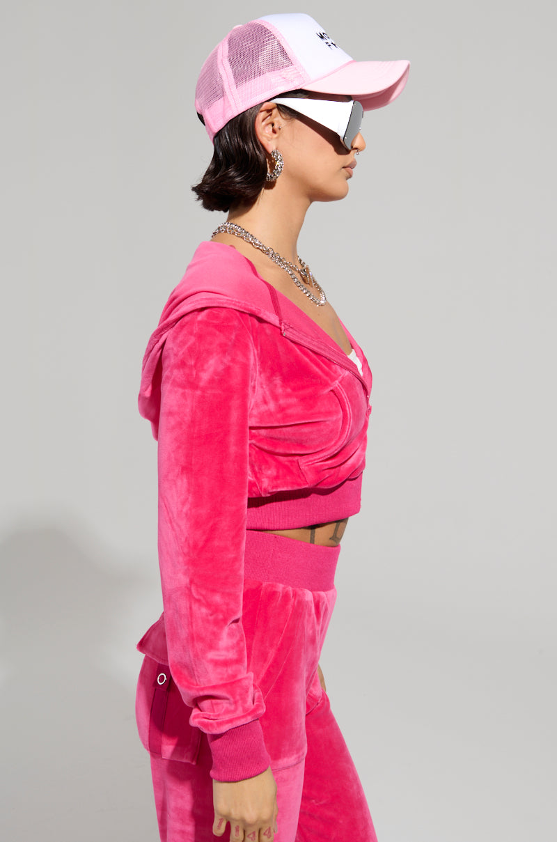 Dressed in the eye-catching VELOUR GALORE FITTED ZIP UP IN PINK, a person exudes true Y2K style with a white and pink baseball cap while looking sideways. Complementing the outfit with oversized sunglasses, hoop earrings, and a necklace set against a plain gray backdrop, they capture retro flair perfectly.