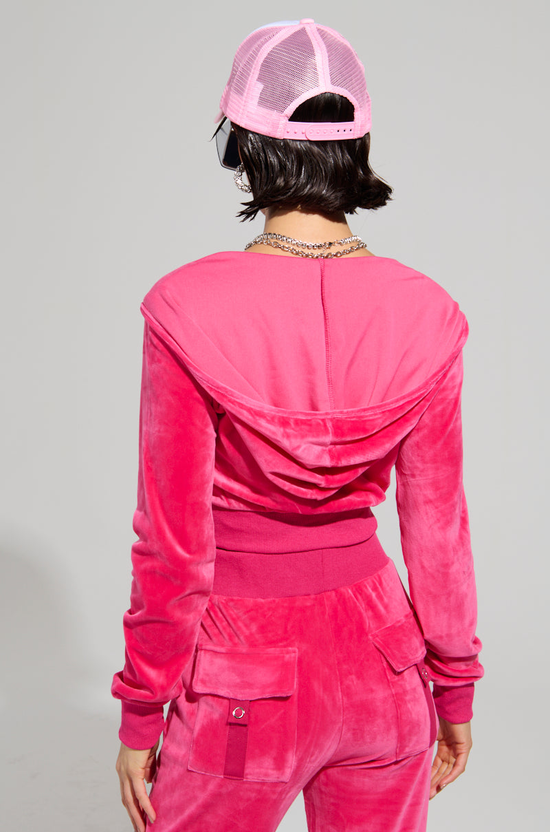 A person wearing the Velour Galore Fitted Zip Up in pink, accompanied by a matching pink mesh-back cap, captures the nostalgic Y2K vibes. The outfit, viewed from behind, includes a hooded sweatshirt and pants with pocket details, complemented by their short dark hair.