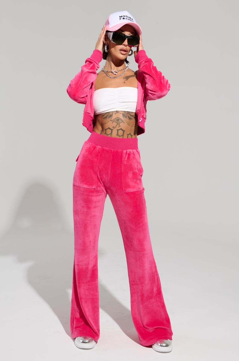 Wearing the bright pink VELOUR GALORE FITTED ZIP UP, this individual exudes Y2K vibes. Paired with a white bandeau top and a pink baseball cap, they confidently pose with hands on their head against a neutral backdrop. Tattoos are visible through the attire, while oversized sunglasses complete the look.