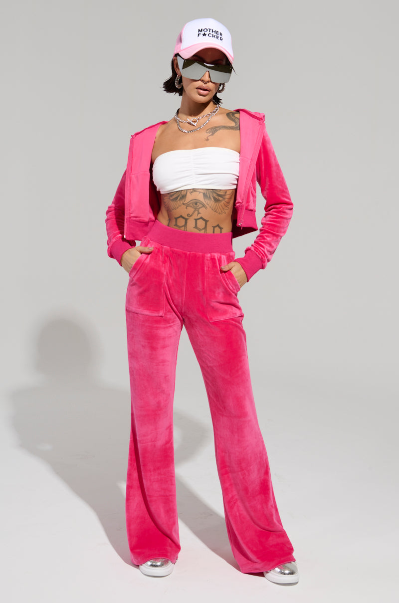 Styled in the Velour Galore Fitted Zip Up in Pink, the individual showcases a velour tracksuit paired with a cropped white tube top and a coordinating pink cap. With sunglasses on and a tattoo adorning their stomach and chest, they casually rest their hands in their pockets, perfectly embodying the Y2K vibe against a simple backdrop.