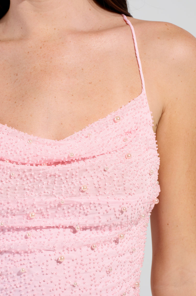 Close-up of a person wearing the LOOK EXPENSIVE COWL NECK EMBELLISHED MINI DRESS, a luxurious pink garment adorned with beads and spaghetti straps. The body-hugging silhouette highlights intricate bead detailing and showcases a softly draped cowl neck. The fair skin tone stands out against the neutral light gray background, conveying an air of elegance.