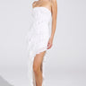 A woman poses in the ELLA MESH TUBE DRESS IN WHITE, featuring cascading ruffles and an asymmetrical hem. She accessorizes with hoop earrings and white strappy heels, her attire elegantly highlighted against a plain backdrop.