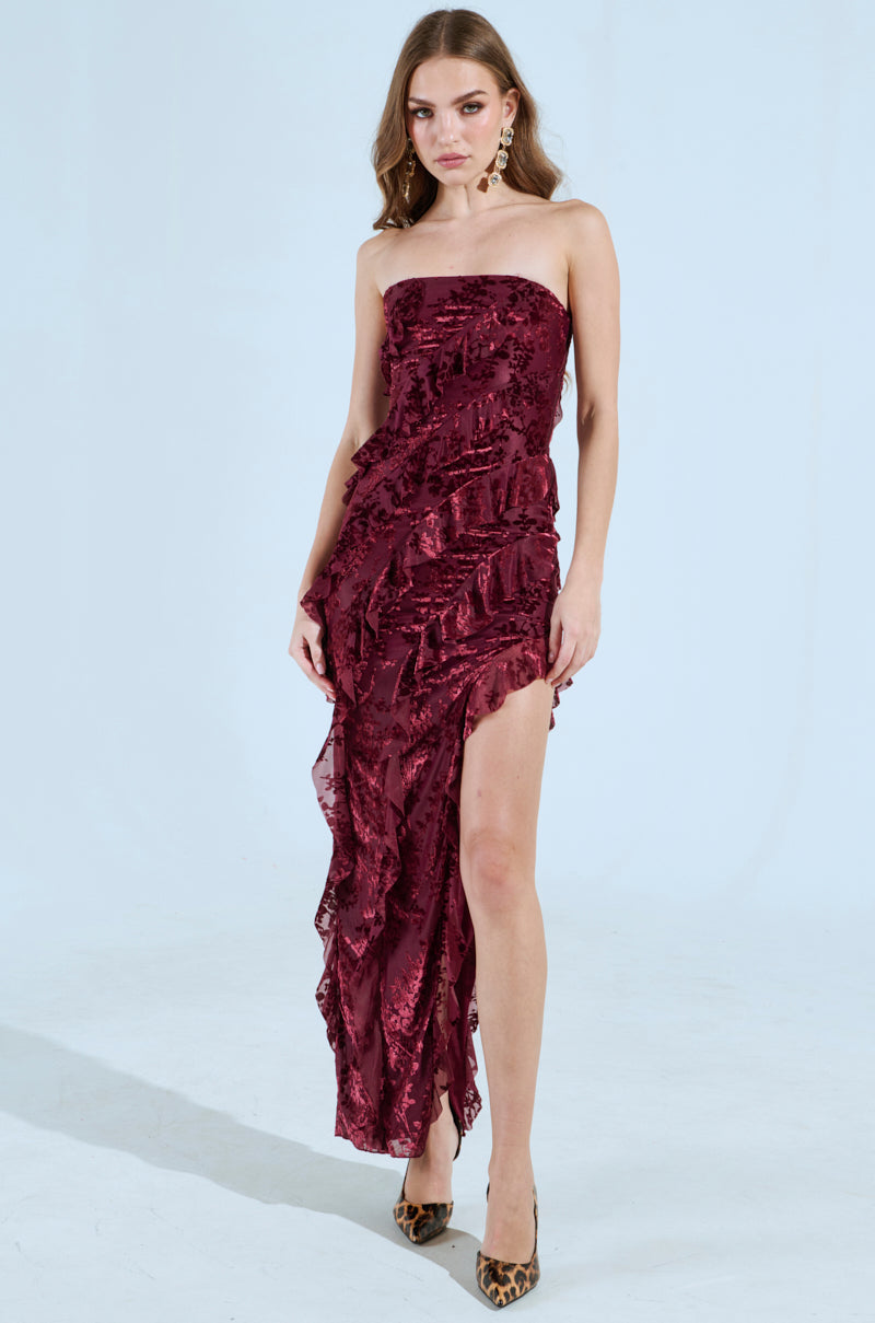 A woman poses in the figure-accentuating CYRUS Velvet Maxi Dress in Burgundy, featuring a strapless design with ruffles and a high slit. She complements her outfit with leopard print high heels against a plain background.