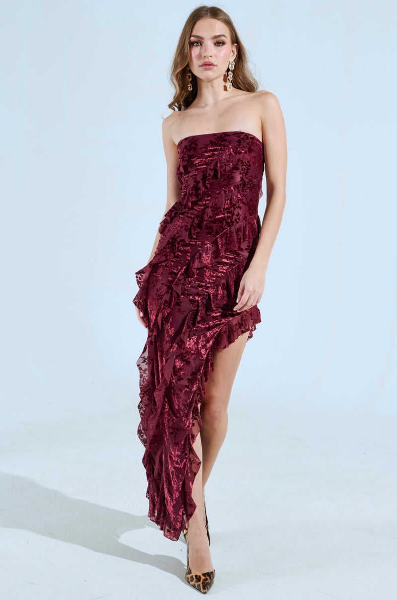 A woman poses in the curve-hugging CYRUS VELVET MAXI DRESS IN BURGUNDY, featuring textured ruffles and an asymmetrical hem. She pairs it with leopard print heels and statement earrings against a plain light background, exuding elegance.
