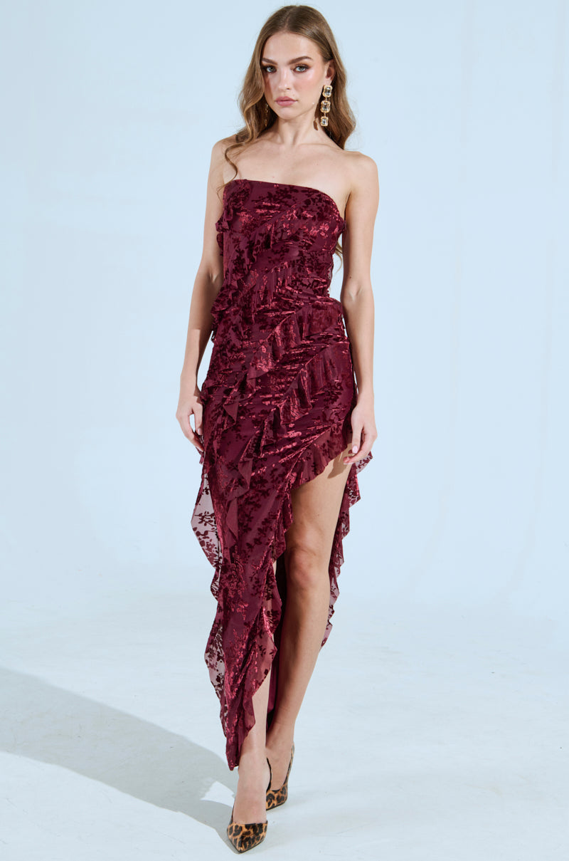 A woman elegantly dons the CYRUS Velvet Maxi Dress in burgundy, featuring a strapless, curve-hugging velvet design with sequin details. Against a plain light background, her long wavy hair and dangling earrings complete her poised and elegant look.