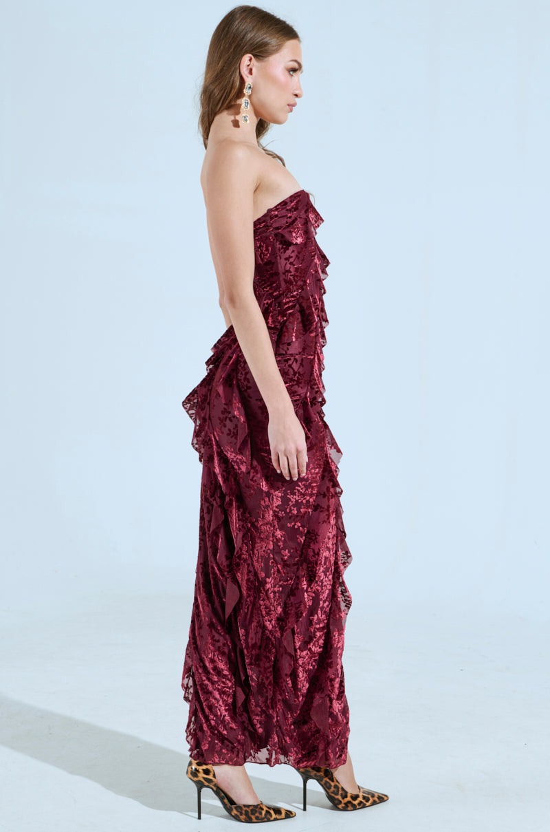 A woman in a strapless, curve-hugging CYRUS VELVET MAXI DRESS IN BURGUNDY poses in profile against a plain backdrop. She accessorizes with statement earrings and leopard-print high heels, and her long hair is styled straight.