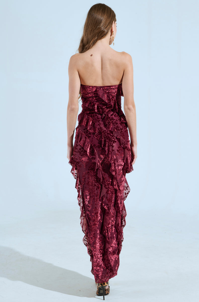A woman with long hair stands facing away, highlighting the CYRUS Velvet Maxi Dress in burgundy, featuring sparkling sequins and layered ruffles. The curve-hugging design is paired with high heels against a plain light background.
