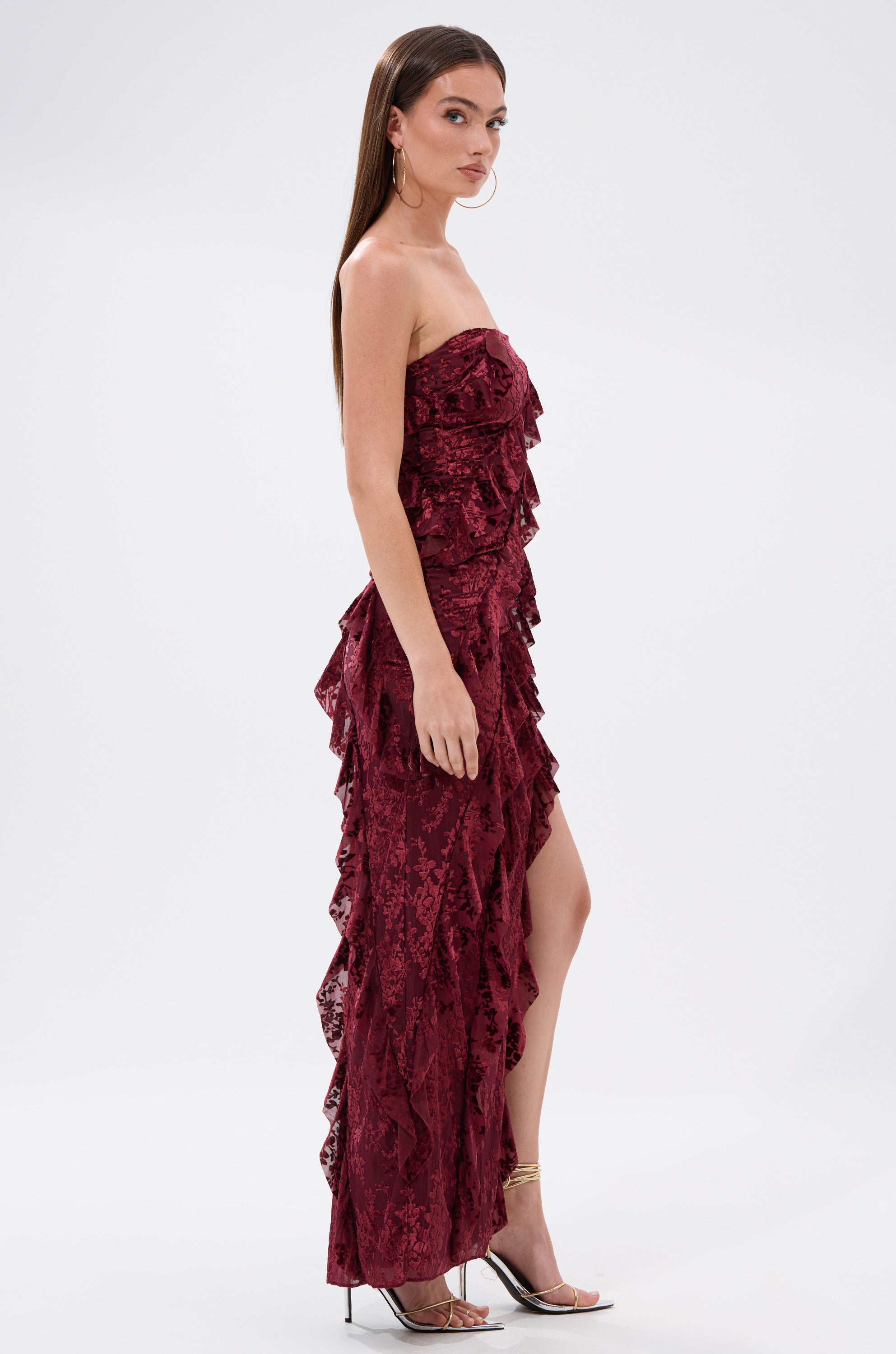A woman in profile wears the WHERE WE GOIN VELVET BURNOUT MIDI DRESS—a strapless burgundy ruffled velvet gown with a thigh-high slit—paired with clear high-heeled sandals, standing against a plain white background.