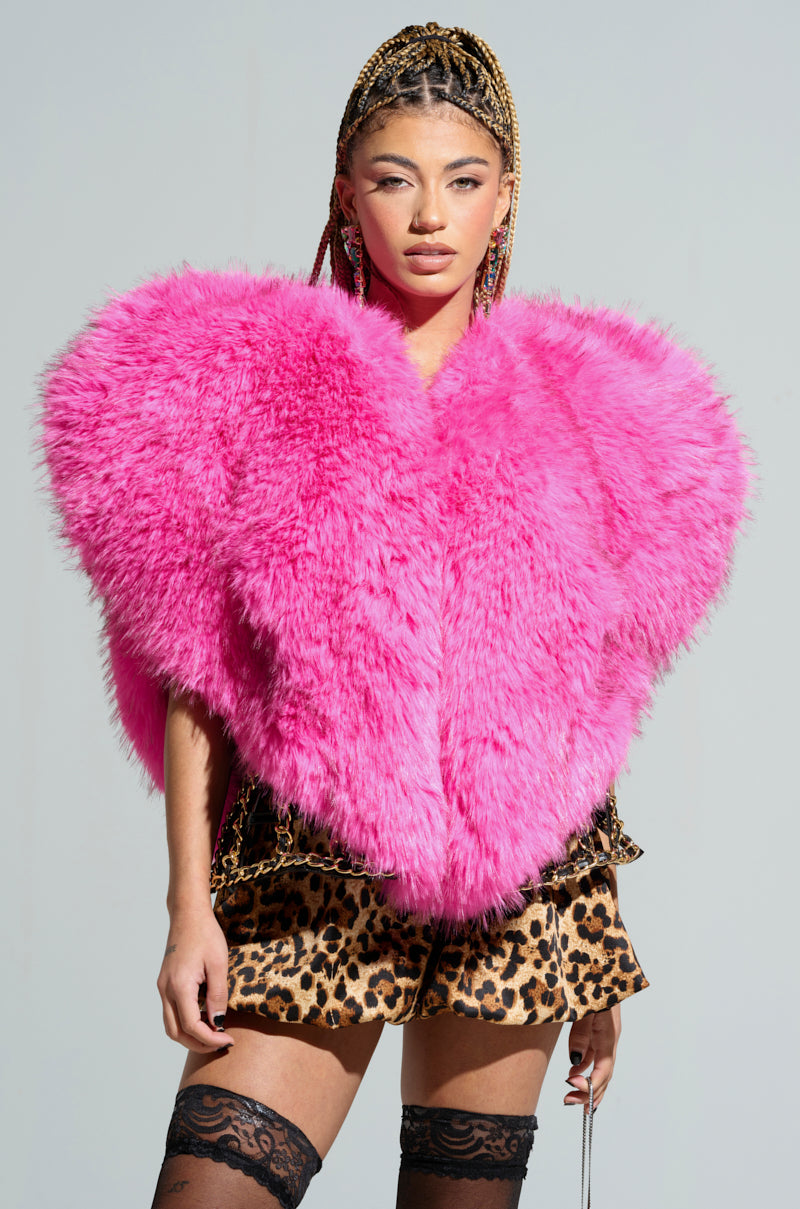 Confidently styled, a person showcases the STAR CROSSED LOVER FAUX FUR HEART VEST, a large and fluffy pink heart-shaped piece, layered over a bold leopard print ensemble. With their hair braided, they pose against a simple gray background.