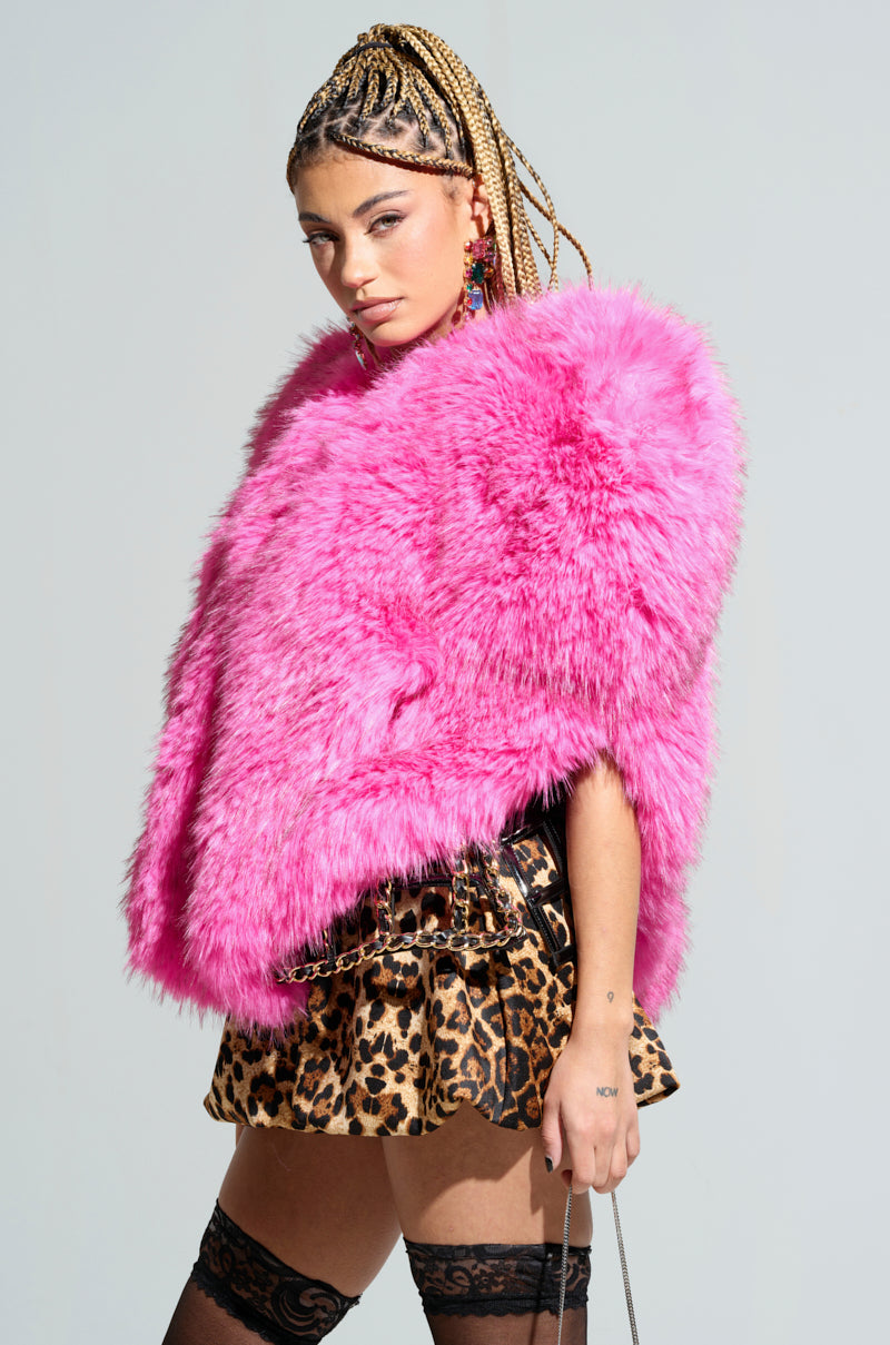 A person with braided hair wears the vibrant STAR CROSSED LOVER FAUX FUR HEART VEST in bright pink, paired with a leopard print skirt. They are accessorized with colorful earrings and stand against a plain background, exuding confidence and style.