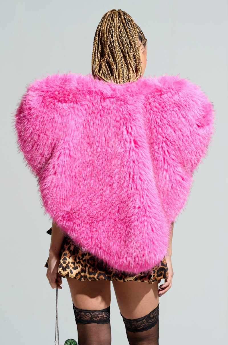 A person with braided hair, draped in the eye-catching STAR CROSSED LOVER faux fur heart vest in pink, paired with a leopard print skirt and black stockings, stands turned away from the camera against a plain background.