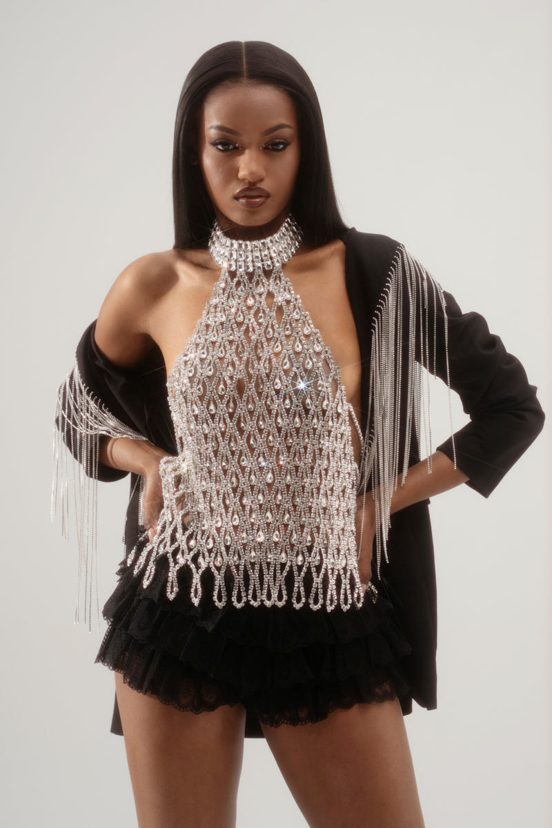 A person confidently poses against a plain background, wearing the LOREN Rhinestone Halter Neck Top in silver, paired with a black jacket featuring fringe sleeves and black ruffled shorts.