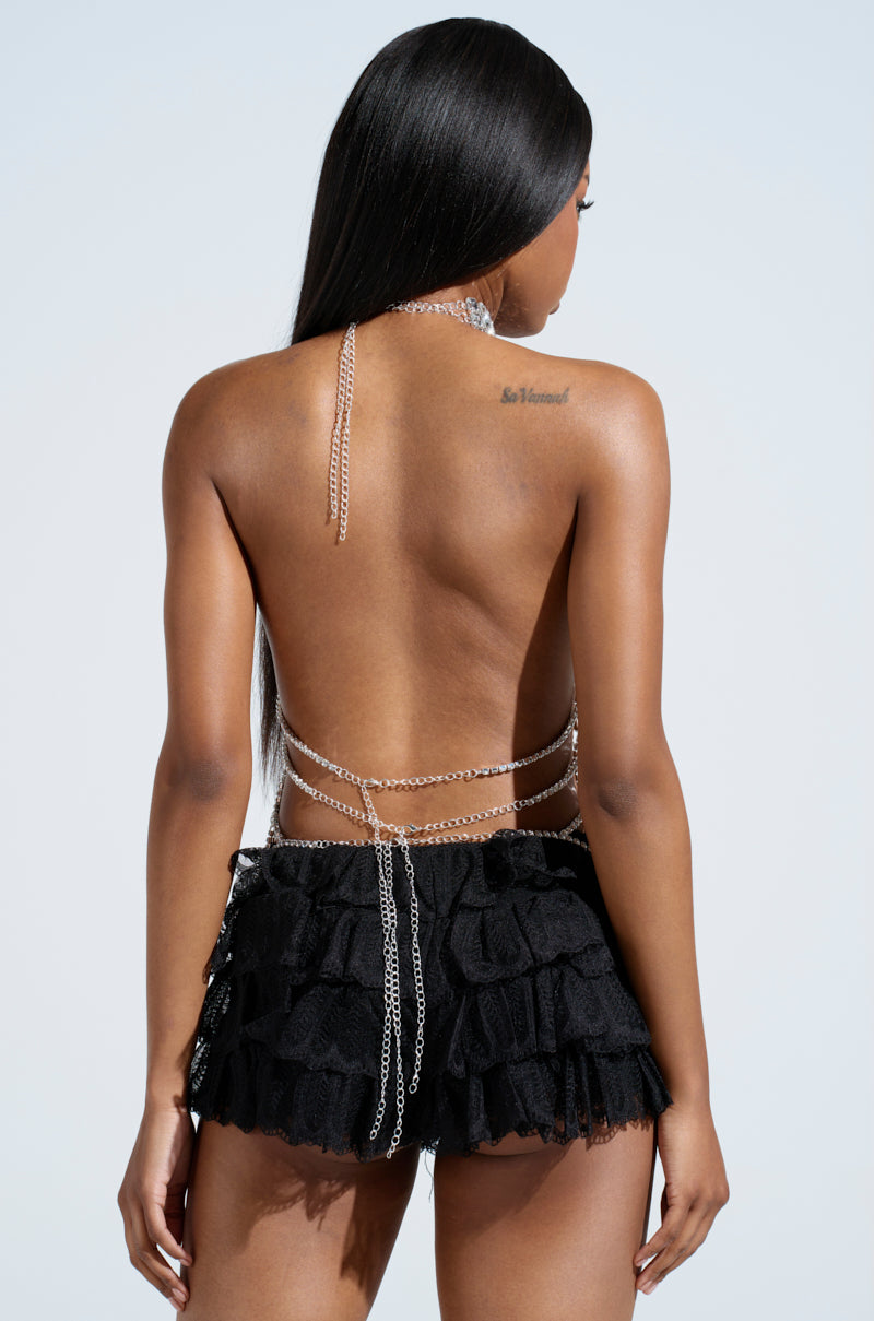 The woman with long black hair faces away, showcasing the Loren Rhinestone Halter Neck Top in silver. The top features backless straps with layered ruffles and cascading rhinestone details, glistening against a simple light blue background.