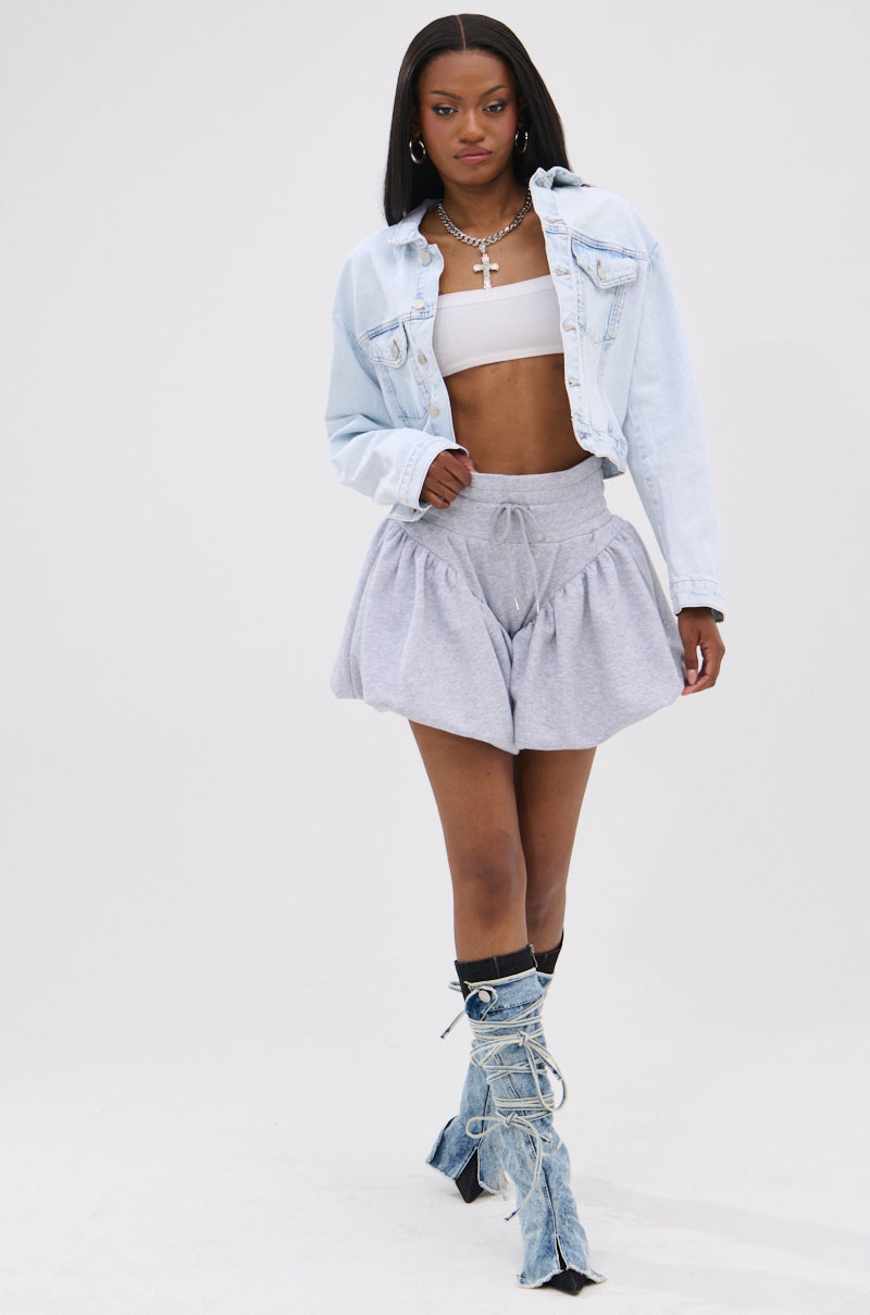 A woman walks confidently in a light blue cotton denim jacket over a white bandeau top, HEATHERED BUBBLE SHORT, layered necklaces, and tall distressed denim heeled boots. The background is plain white.