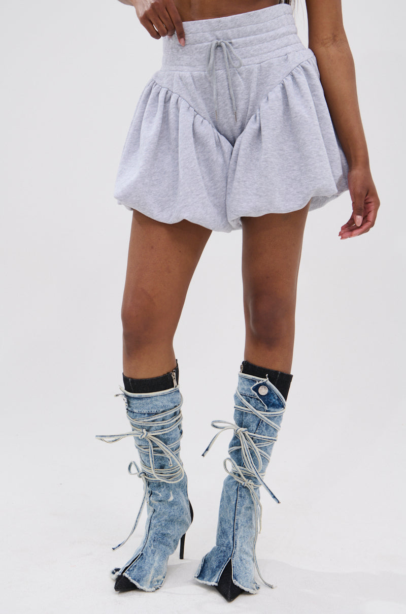 Wearing HEATHERED BUBBLE SHORTS in light gray, high-rise cotton, and knee-high lace-up denim boots with frayed edges, a person stands against a plain white background.