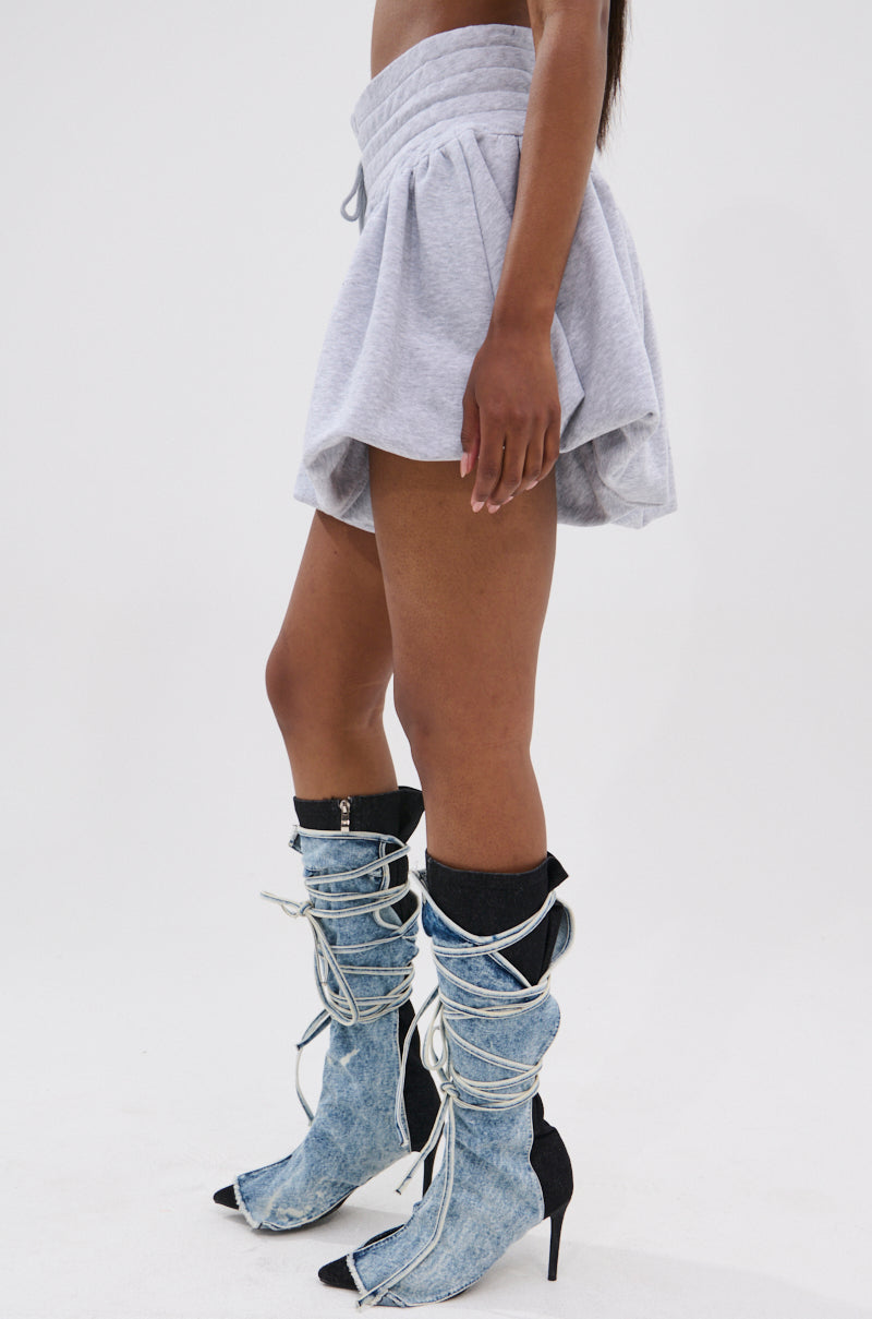 A person stands sideways against a white background wearing the HEATHERED BUBBLE SHORT in light gray and blue denim high-heeled boots with white laces. Their head is not visible in the image.