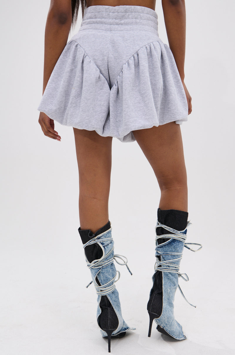 A person stands facing away, wearing the HEATHERED BUBBLE SHORT in light gray cotton with a high-rise fit and two-tone denim high-heeled boots featuring white laces, set against a plain white background.