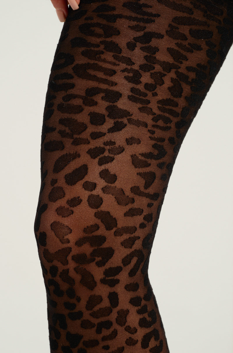 Close-up of a leg adorned with sheer black CHEETAH SISTA TIGHTS featuring a leopard print pattern crafted from a stretchy nylon blend, set against a light and plain background.