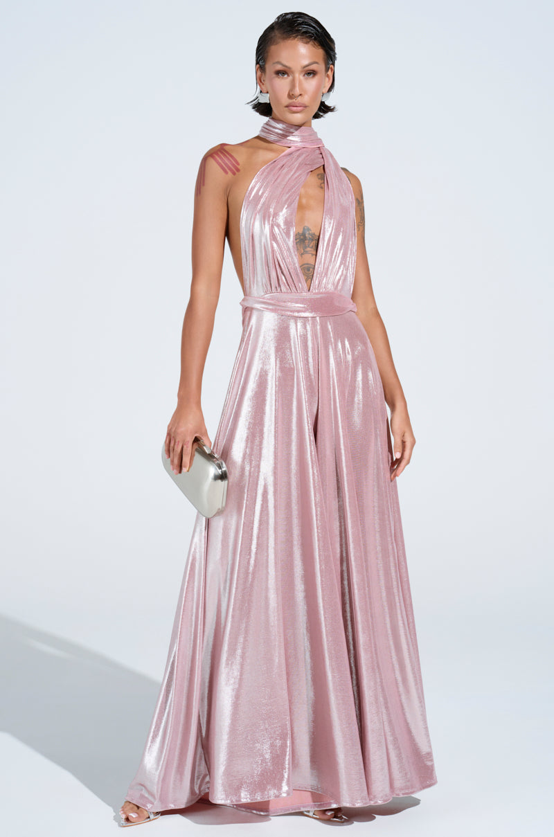 A person poses in a dazzling pink SHINE BRIGHT WEAR MULTIPLE WAYS JUMPSUIT featuring a deep V neckline and flowing floor-length skirt. They hold a silver clutch, their hair styled in a wet look, against a plain light background.