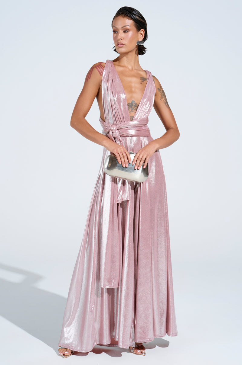 Wearing the SHINE BRIGHT WEAR MULTIPLE WAYS JUMPSUIT in pastel pink metallic with a plunging neckline and self-tie bodice, they pair it with a silver clutch. Tattoos adorn their arm and chest, while their slicked-back hair complements their confident pose against a light background.