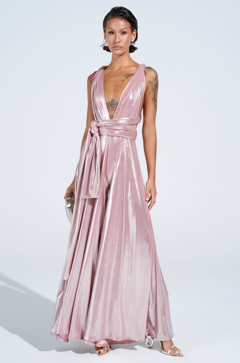 A person in the SHINE BRIGHT WEAR MULTIPLE WAYS JUMPSUIT, a sleeveless pink satin piece with a self-tie bodice and wide-leg pants, stands confidently against a plain background. With short hair, visible arm tattoos, and open-toed heels, they hold a small silver clutch.