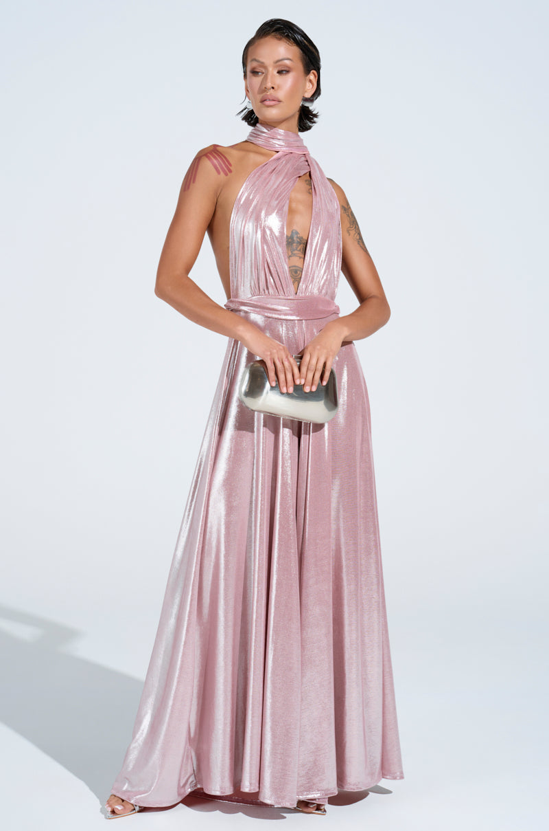 A person is posing in the SHINE BRIGHT WEAR MULTIPLE WAYS JUMPSUIT—a metallic pink piece featuring a self-tie bodice and plunging halter neckline, designed with a crisscross pattern. They're holding a silver clutch, and their sleek hair is styled back against a simple, light background.