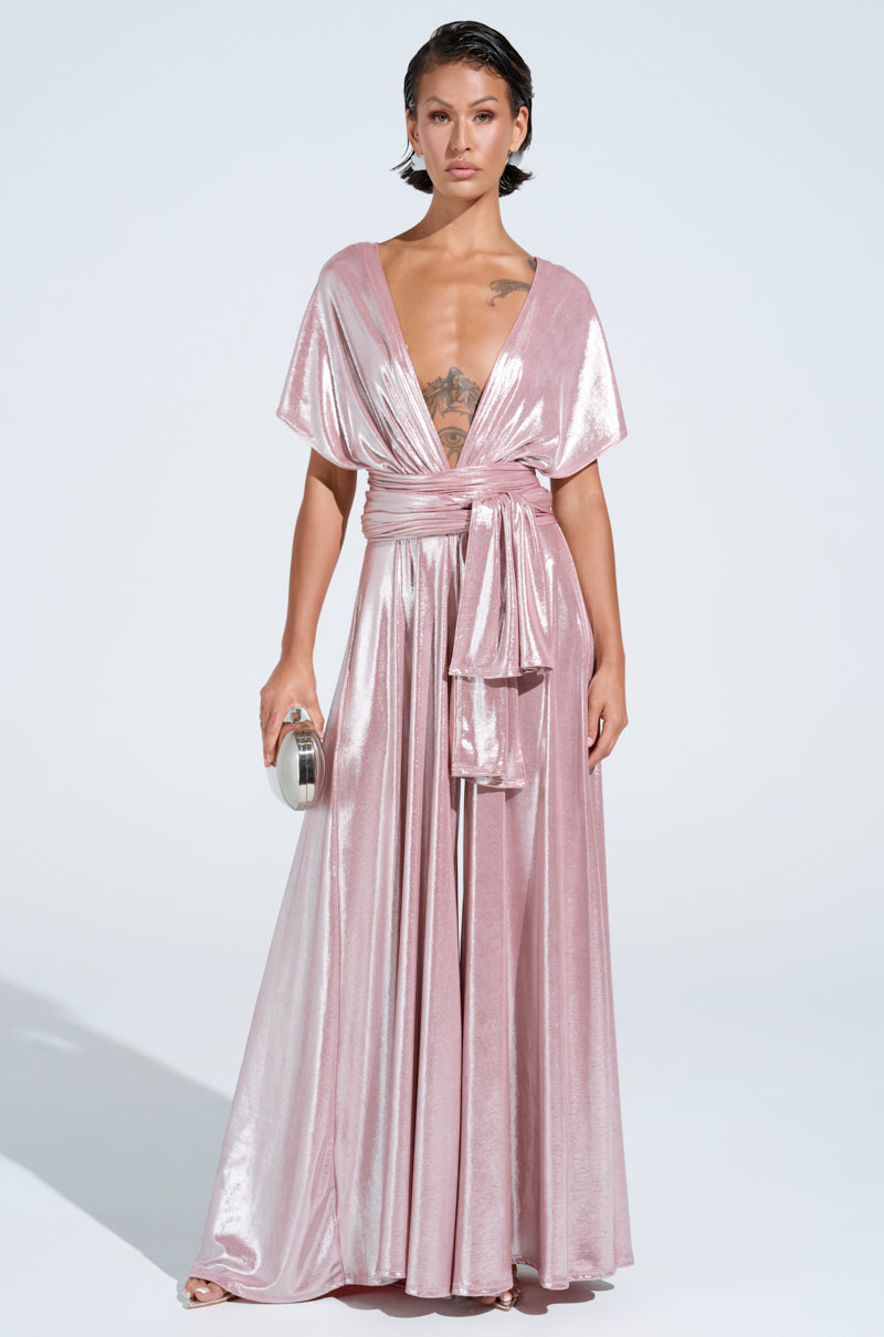 Person in the SHINE BRIGHT WEAR MULTIPLE WAYS JUMPSUIT, a flowing metallic pink design with a deep neckline and self-tie bodice, holding a small silver clutch. They pose confidently on a light background, exuding elegance.