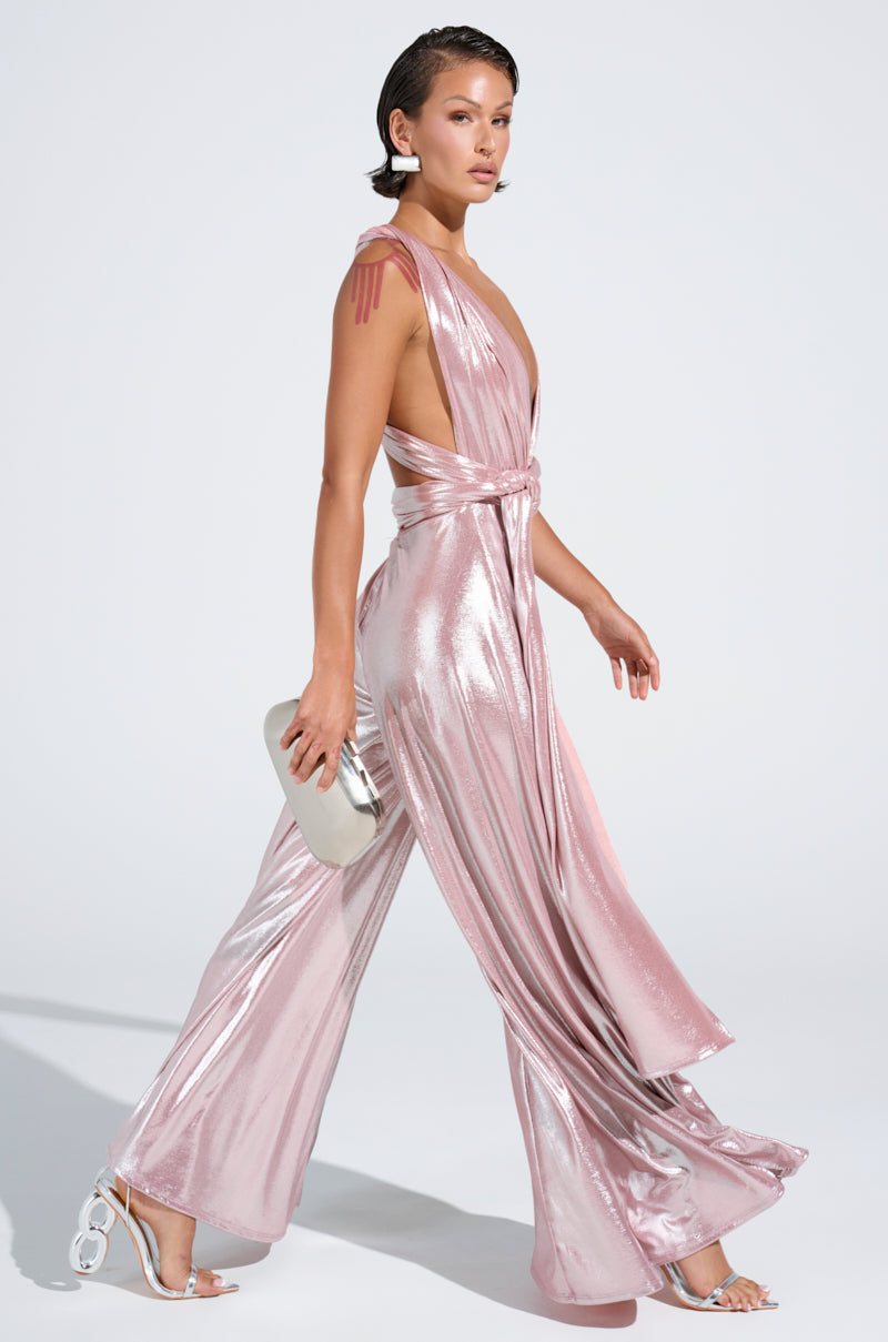 A person with short hair strides confidently in the SHINE BRIGHT WEAR MULTIPLE WAYS JUMPSUIT, a shimmering pink metallic piece with a deep neckline, self-tie bodice, and wide legs. They hold a silver clutch and wear white heels against a plain background that emphasizes the elegant ensemble.