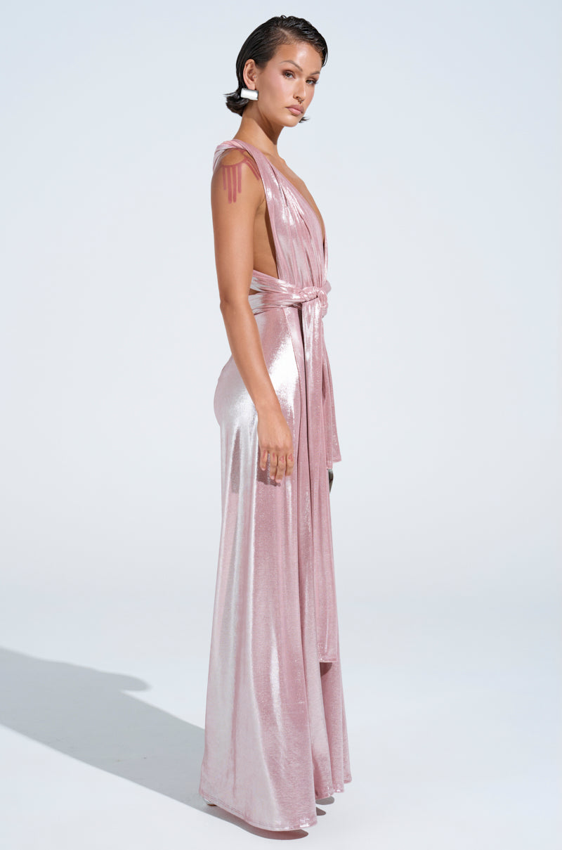 A woman in the SHINE BRIGHT WEAR MULTIPLE WAYS JUMPSUIT poses sideways against a bright background. The shimmering pink gown, with its self-tie bodice, elegantly compliments her short hair and white earrings, exuding effortless charm.